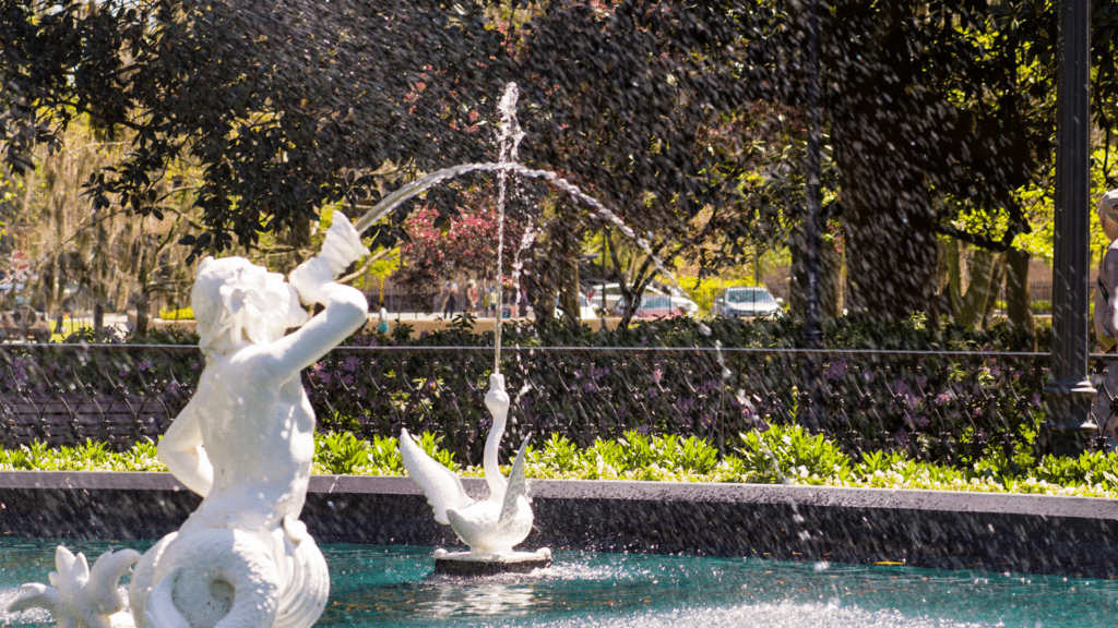White fountain sculptures spray water into a pool surrounded by trees and greenery.