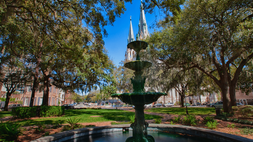 Tiered fountain beneath leafy trees, with twin church steeples rising behind a Savannah square.