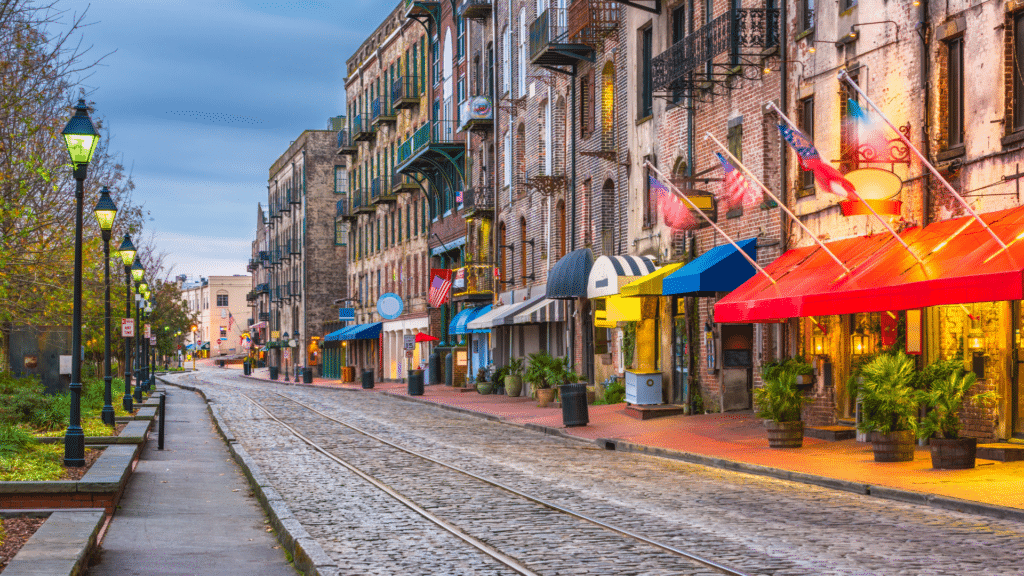 Colorful storefront awnings line Savannah’s cobblestone River Street beside embedded rail tracks.