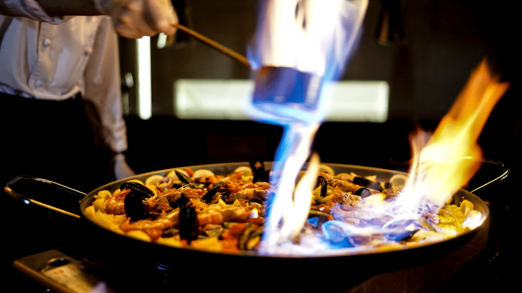 Chef tending a pan of seafood paella as flames rise above the cooking surface.