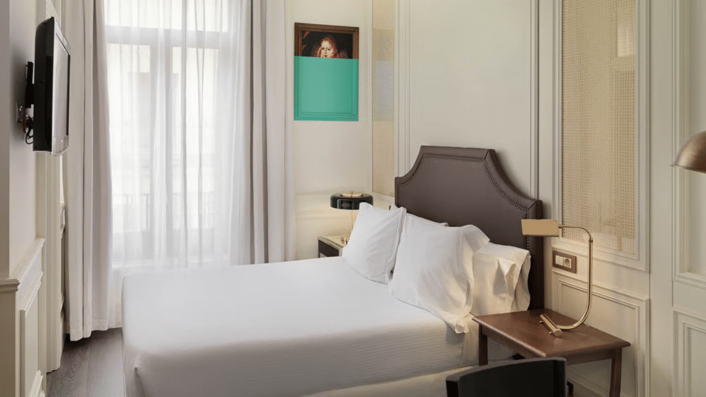 Bright hotel room with a double bed, white linens, a desk, and tall curtained windows.