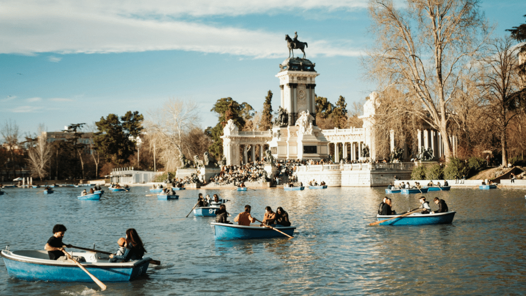 Visitors row blue boats across Retiro Park’s lake in front of the Alfonso XII monument.
